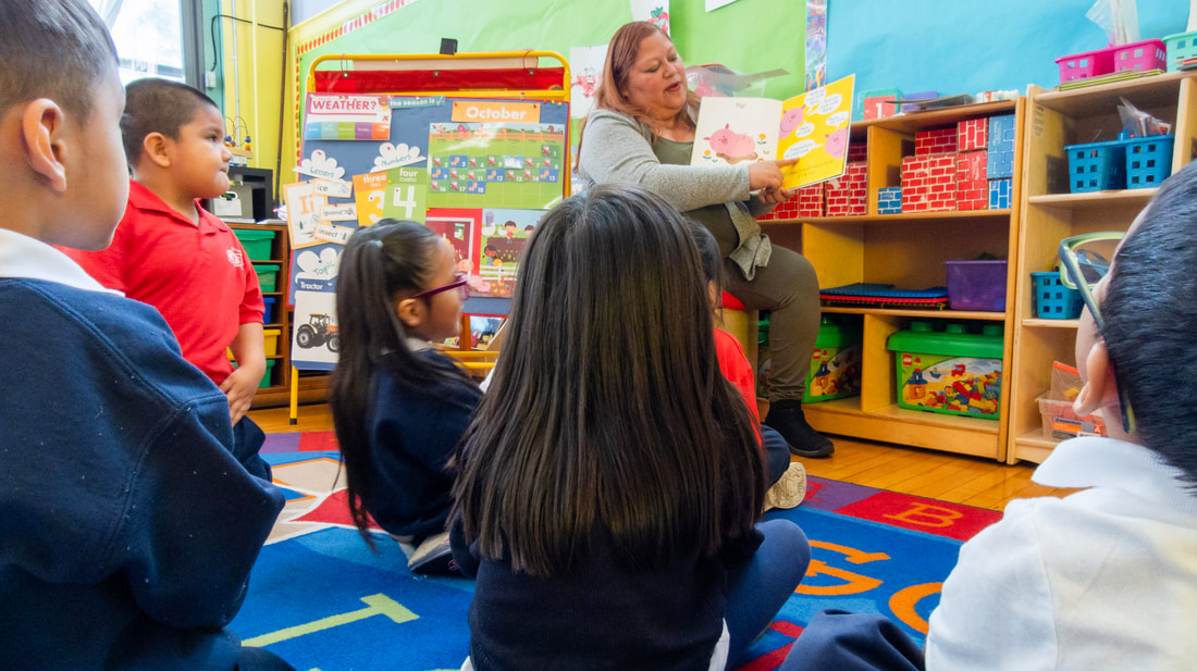 La Sra. Gutiérrez, profesora de Preescolar 3 y 4, está sentada delante de la clase leyendo un libro en voz alta. Los alumnos se sientan en una alfombra de colores brillantes frente a ella.