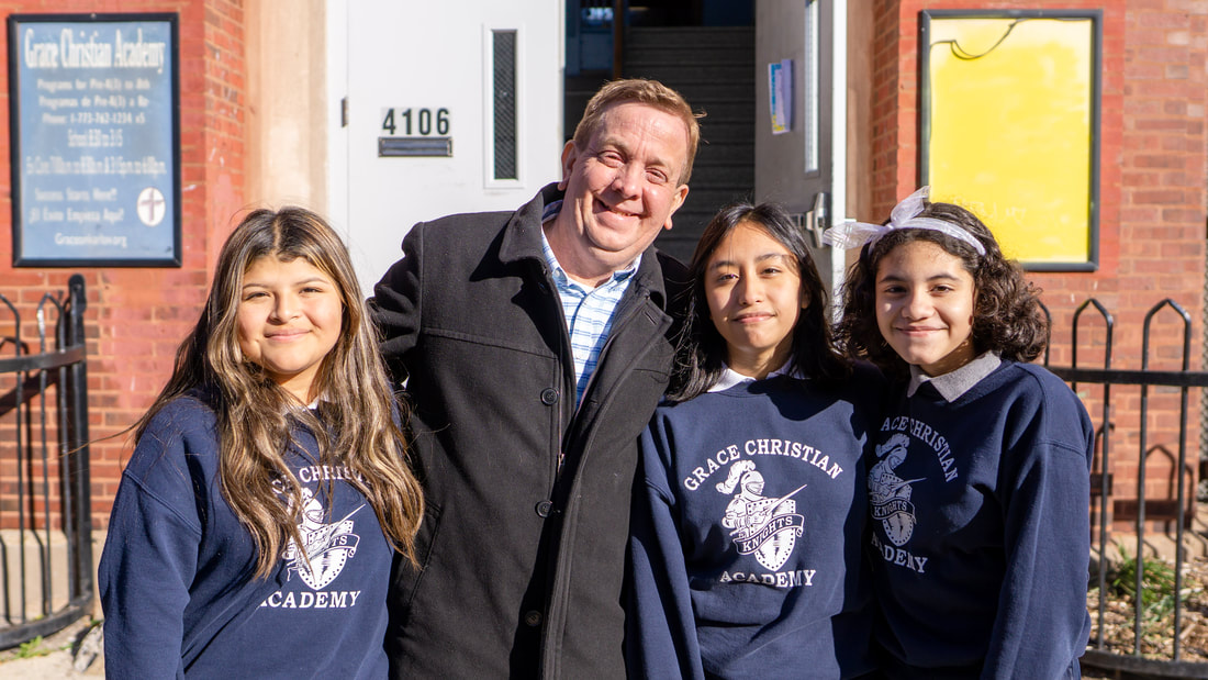 El director Carlo Giannotta de Grace Christian Academy sonriendo con tres alumnas de secundaria en un día soleado frente a la escuela.