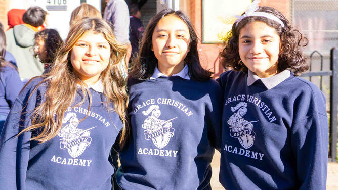 Tres alumnas de secundaria sonríen abrazadas en un día soleado frente a la escuela. Todas llevan sudaderas azules de Grace Christian Academy.