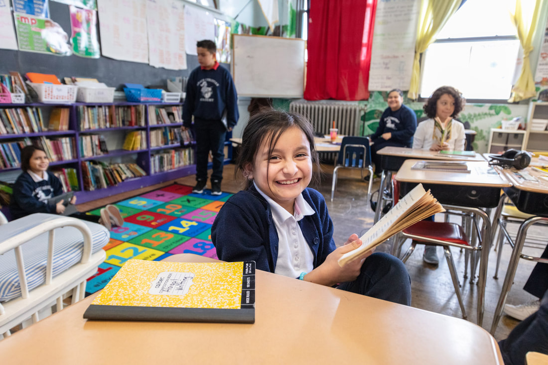 Una estudiante de primaria sentada en su pupitre, con un libro en la mano y sonriendo a la cámara, mientras cuatro alumnos están de pie y sentados al fondo.